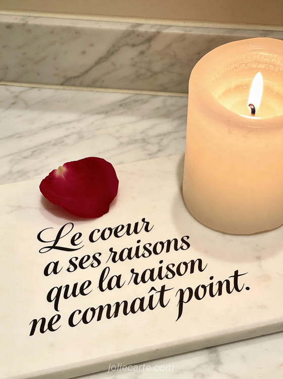 Cœur composé de pétales de roses rouges sur marbre blanc avec lumière de bougie et le texte Le cœur a ses raisons que la raison ne connaît point