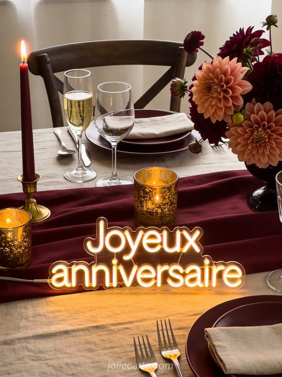 Texte Joyeux anniversaire en lettres lumineuses chaudes sur fond bordeaux avec verres de champagne, bougies dorées et bouquet de dahlias
