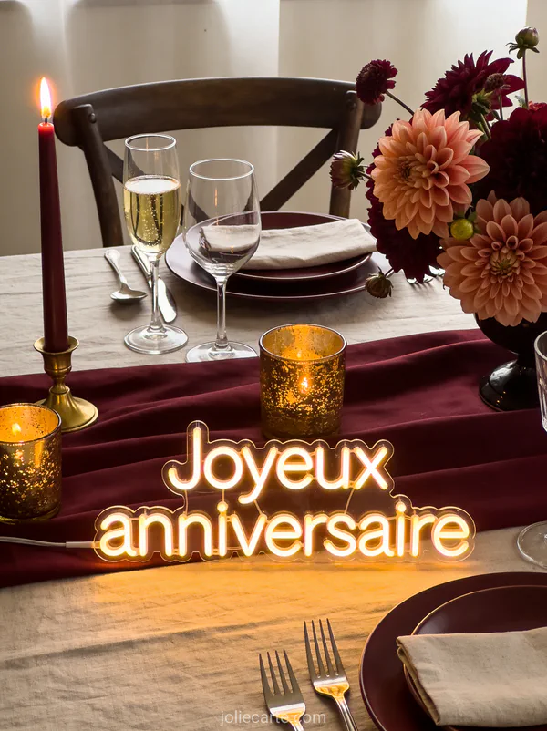 Texte Joyeux anniversaire en lettres lumineuses chaudes sur fond bordeaux avec verres de champagne, bougies dorées et bouquet de dahlias
