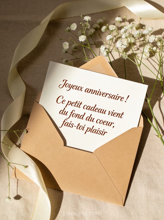 Enveloppe kraft ouverte sur nappe en lin beige avec brins de gypsophile et ruban satin crème, lumière douce latérale avec texte anniversaire manuscrit