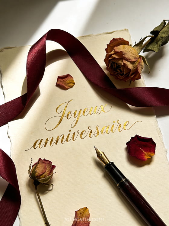 Calligraphie à la plume dorée sur parchemin crème entouré de fleurs séchées et ruban bordeaux avec le texte Joyeux anniversaire