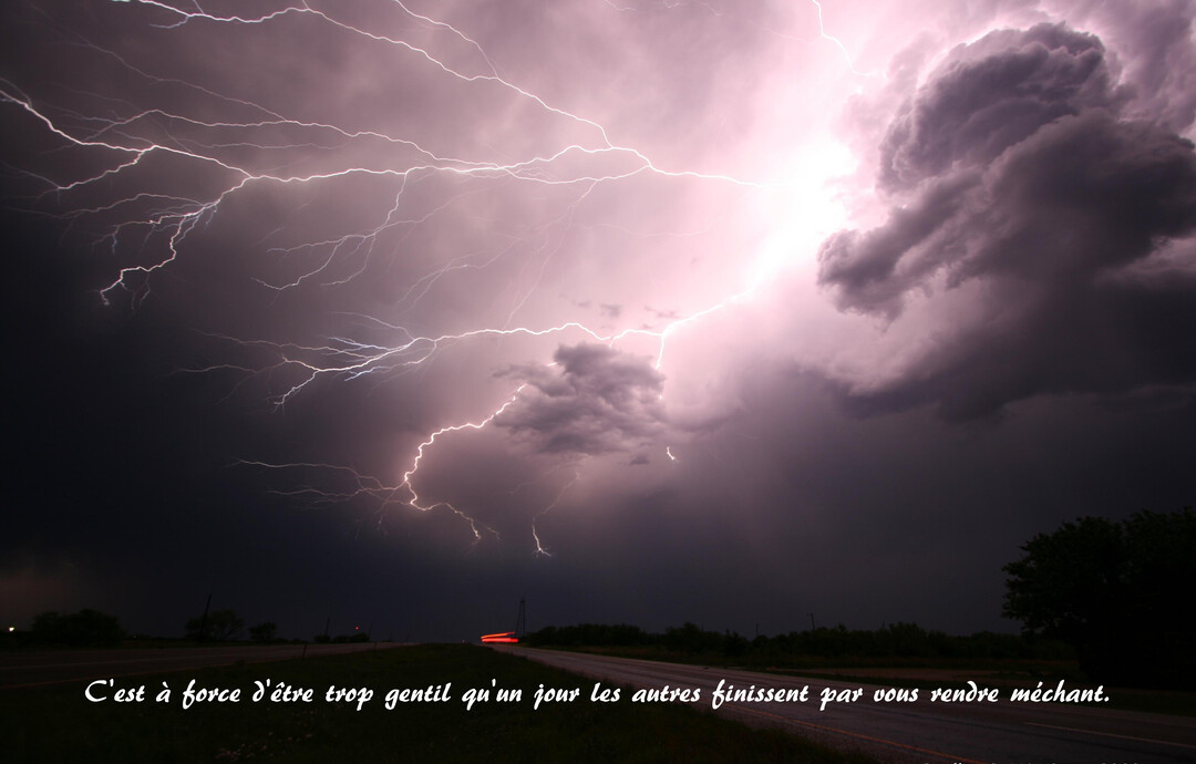Photographie nocturne d'un violent orage, où de multiples éclairs blancs et violets illuminent un ciel sombre au-dessus d'une route déserte.