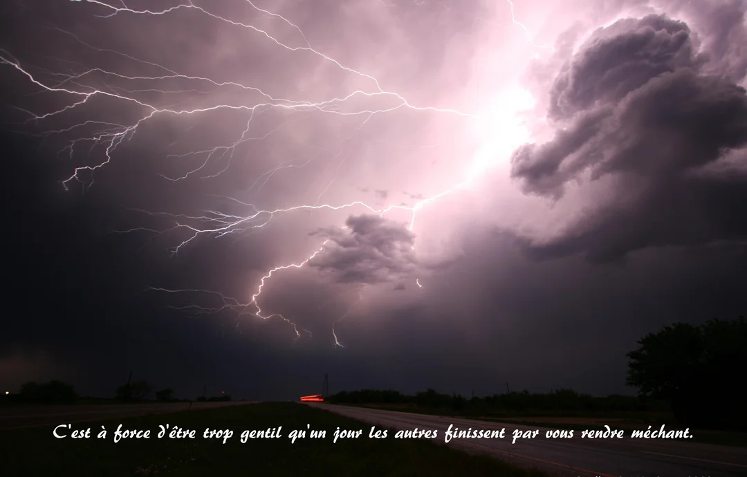 Photographie nocturne d'un violent orage, où de multiples éclairs blancs et violets illuminent un ciel sombre au-dessus d'une route déserte.