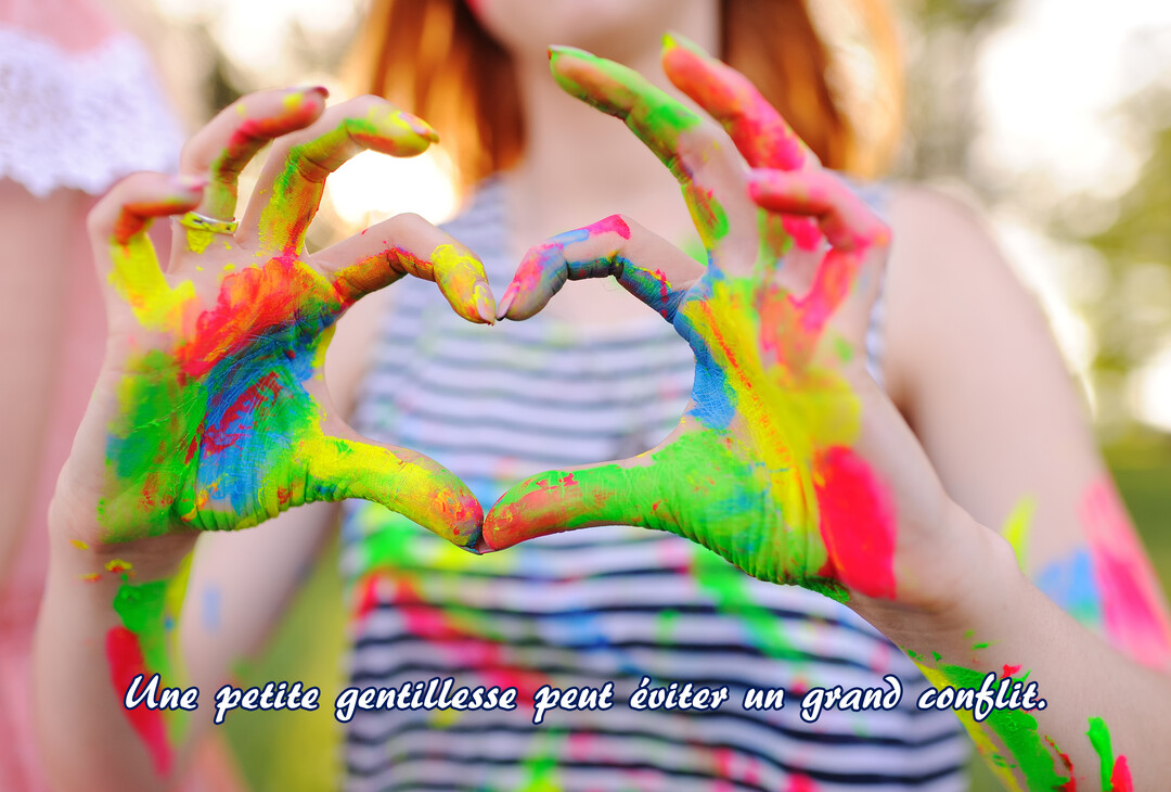 Des mains couvertes de taches de peinture vives et colorées sont jointes pour former un cœur, avec en arrière-plan flou une personne.