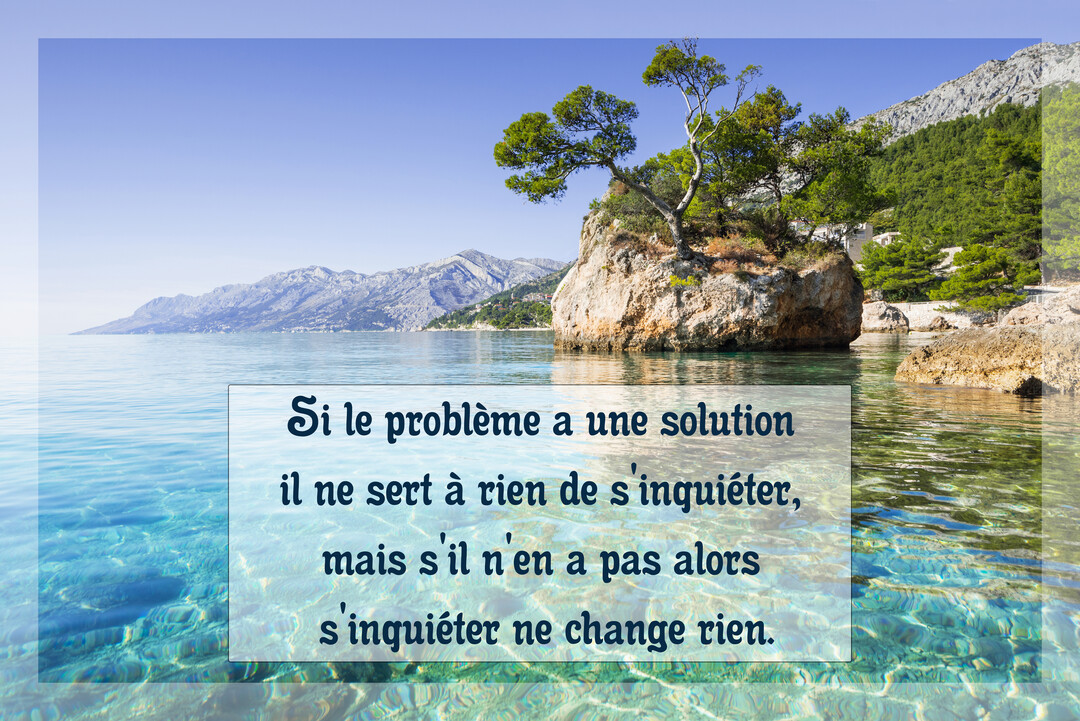 Paysage estival d'une crique à l'eau cristalline, avec un rocher surmonté d'un arbre et des montagnes se découpant sur un ciel bleu.