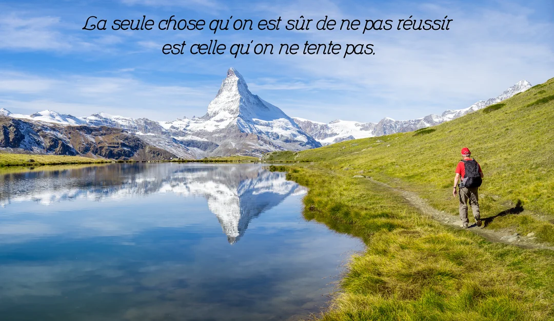 Un homme randonne le long d'un lac de montagne sous un ciel bleu, avec le pic enneigé du Cervin se reflétant dans l'eau.