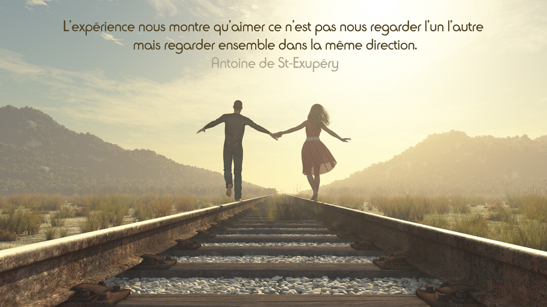 Un couple vu de dos se tient la main et marche en équilibre sur une voie ferrée, s'avançant vers un horizon lumineux et montagneux.