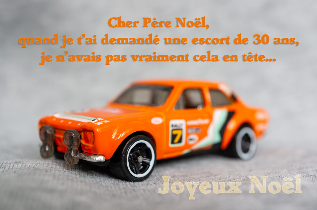 Une carte de vœux de Noël humoristique présente une voiture de rallye miniature orange sur une surface grise, avec un texte amusant.