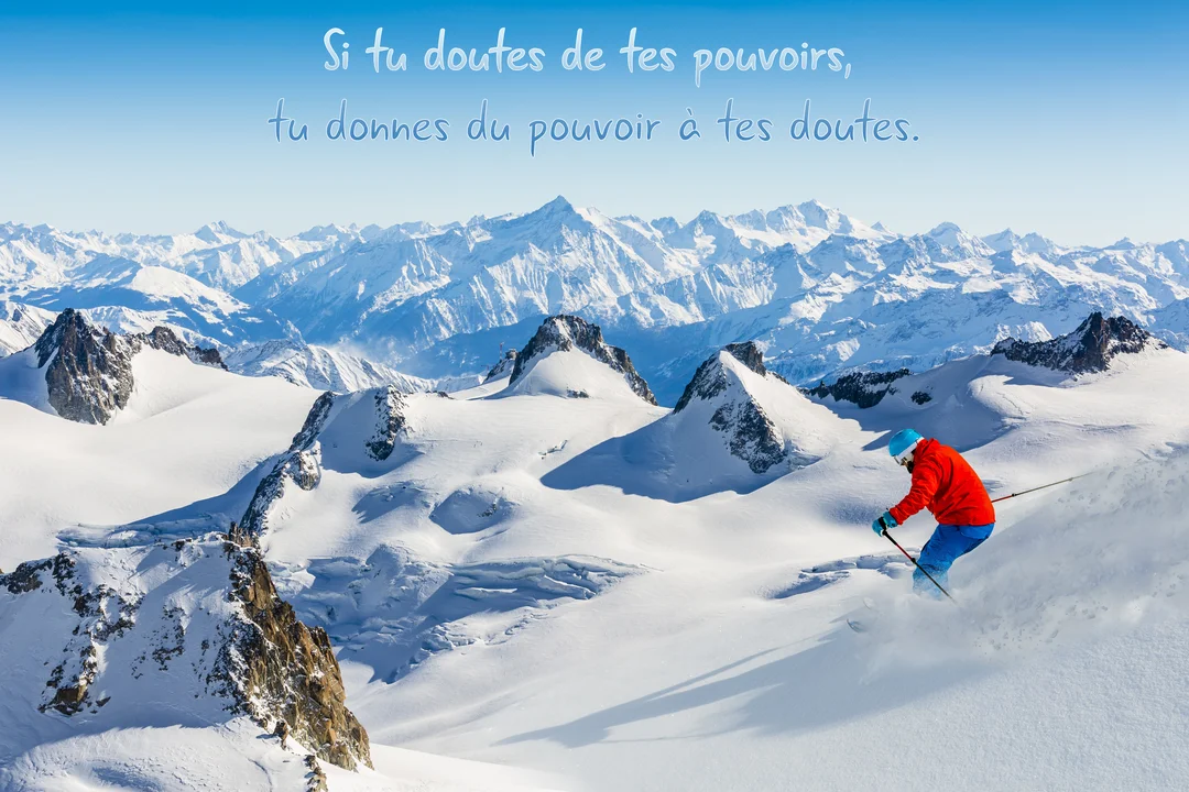 Un skieur en tenue rouge descend une pente de neige poudreuse, avec en arrière-plan un panorama de hautes montagnes sous un ciel bleu.