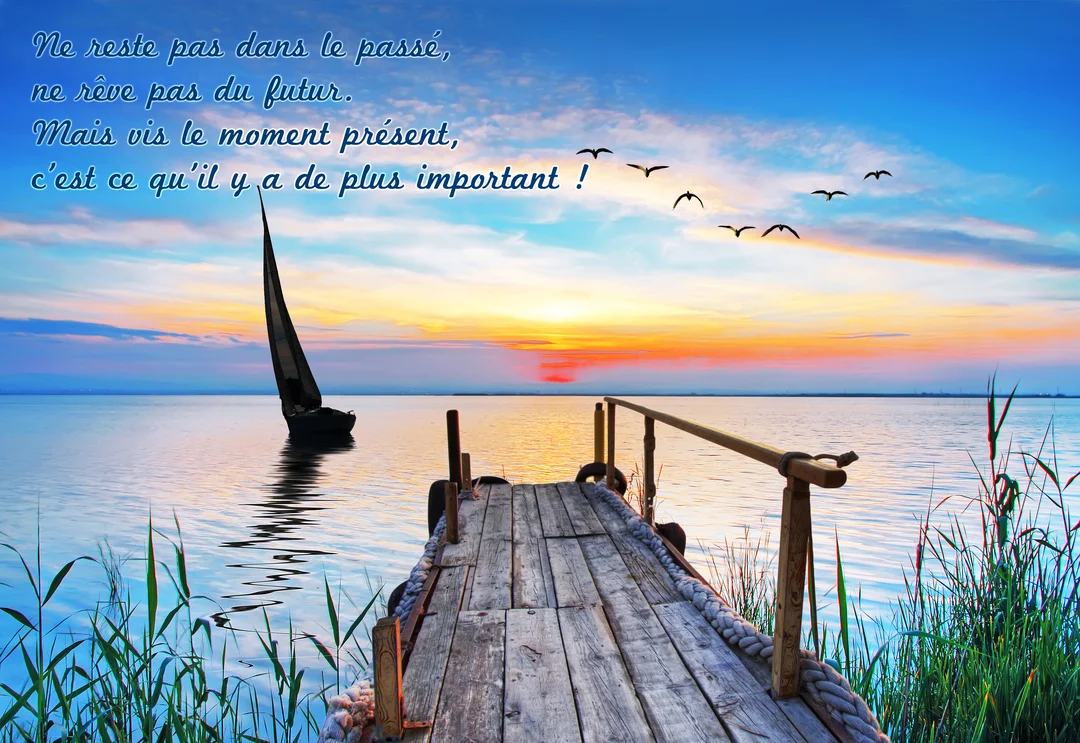 Un ponton en bois mène vers une étendue d'eau calme au coucher du soleil, où navigue un voilier sous un ciel coloré.