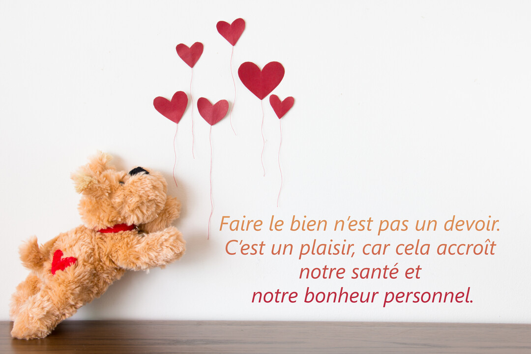 Un ourson en peluche marron est allongé sur une surface en bois, semblant poursuivre des ballons en forme de cœur rouges sur un fond blanc.