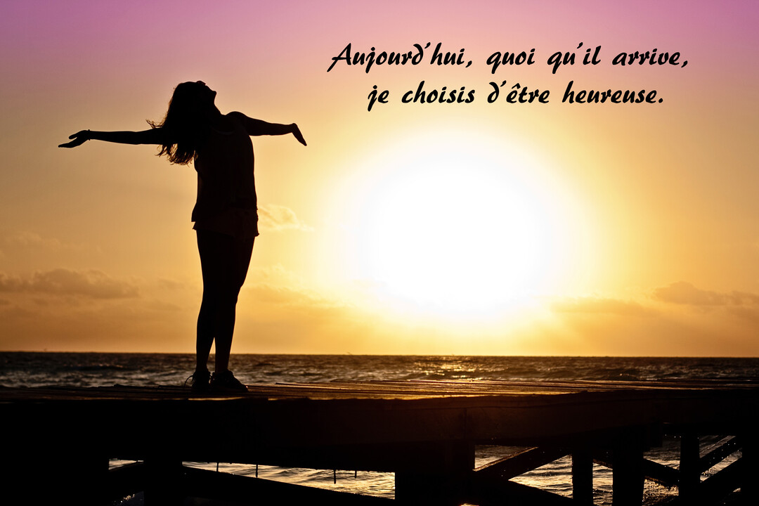 En contre-jour, la silhouette d'une femme se tient sur un ponton, les bras écartés face à un soleil éclatant au-dessus de la mer.