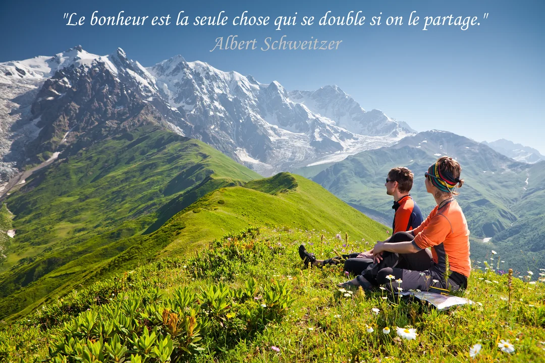 Un homme et une femme sont assis dans une prairie en montagne, contemplant une vaste chaîne de sommets enneigés sous un ciel d'été.