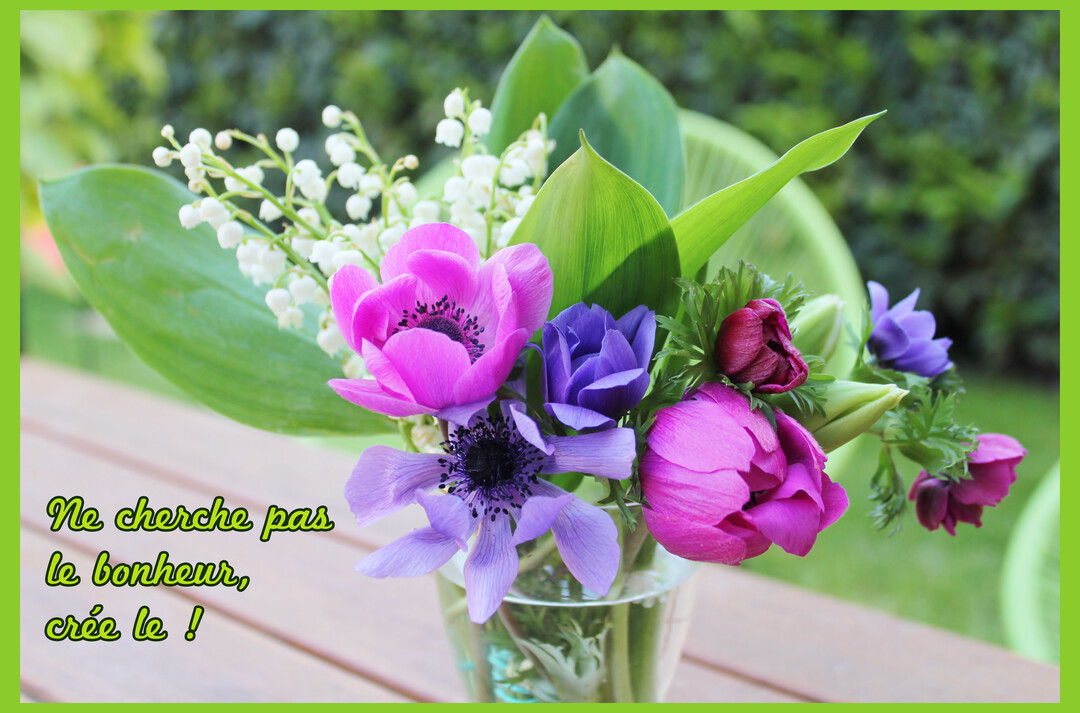 Un bouquet de fleurs printanières, avec des anémones roses et violettes et du muguet blanc, est posé dans un vase en verre sur une table en bois.