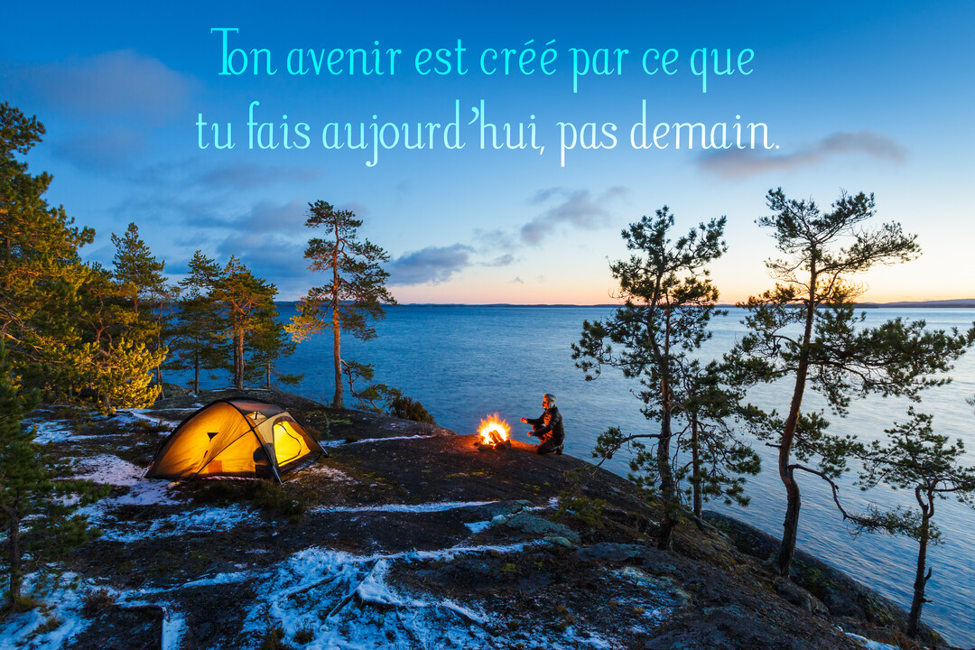 Au crépuscule, une personne est assise près d'un feu de camp et d'une tente illuminée, au bord d'un lac dans un paysage enneigé.