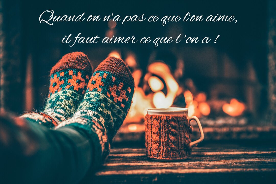 En premier plan, des pieds chaussés de grosses chaussettes en laine sont posés près d'une tasse, devant un feu de cheminée flou.