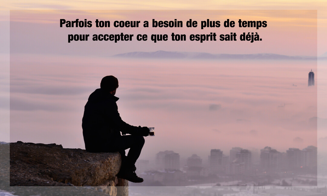 Un homme en silhouette est assis sur un rocher et contemple une ville recouverte par une épaisse brume sous un ciel crépusculaire.