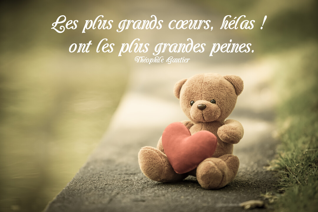 Un petit ours en peluche marron est assis sur une surface en pierre, tenant un cœur en tissu rouge, sur un arrière-plan flou.