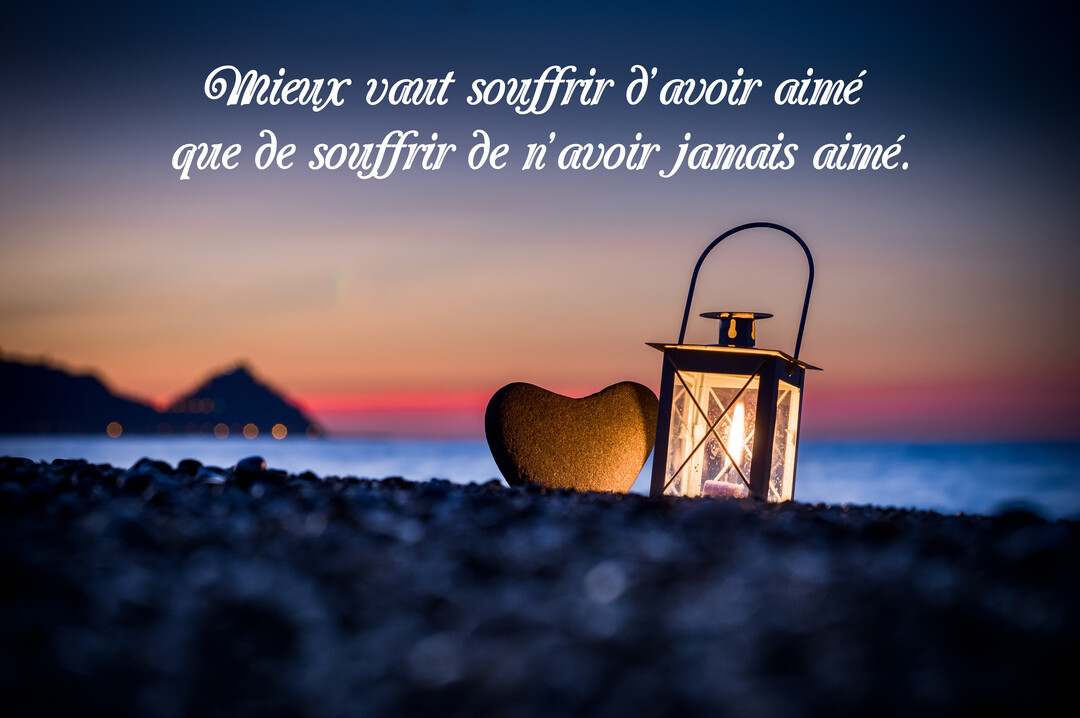 Au premier plan, une lanterne allumée et un galet en cœur sur une plage de galets, avec la mer et un coucher de soleil en arrière-plan.