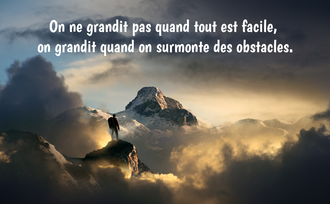 Un homme avec un sac à dos se tient sur un pic rocheux, dominant une mer de nuages et une chaîne de montagnes enneigées.