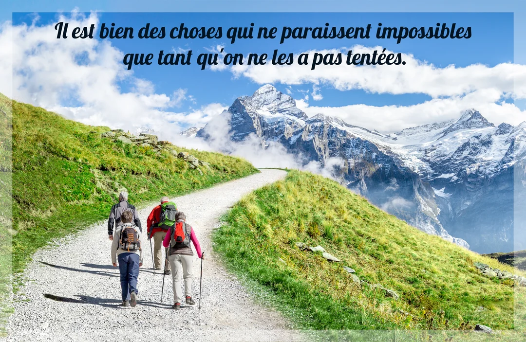 Un groupe de quatre randonneurs équipés avance sur un sentier de montagne, s'élevant vers un sommet enneigé et majestueux sous un ciel bleu.