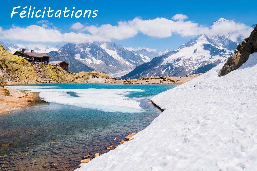 Paysage ensoleillé de haute montagne avec des sommets enneigés, un lac turquoise partiellement gelé, et un chalet sur un promontoire rocheux.