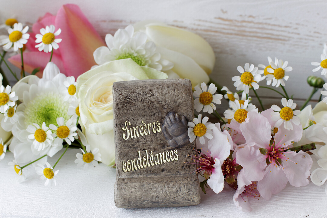 Une plaque en pierre gravée 'Sincères condoléances' est posée au milieu d'un bouquet de fleurs blanches et roses sur un fond en bois.