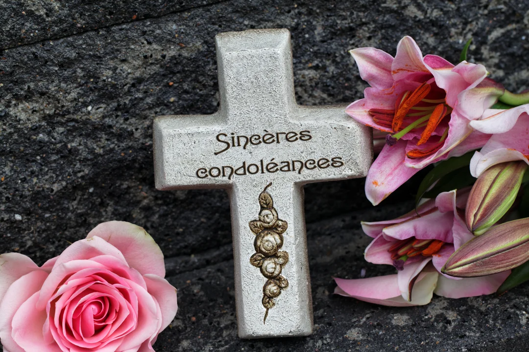 Une croix en pierre gravée de 'Sincères condoléances' est posée sur une roche sombre, entourée d'une rose et de lys roses.