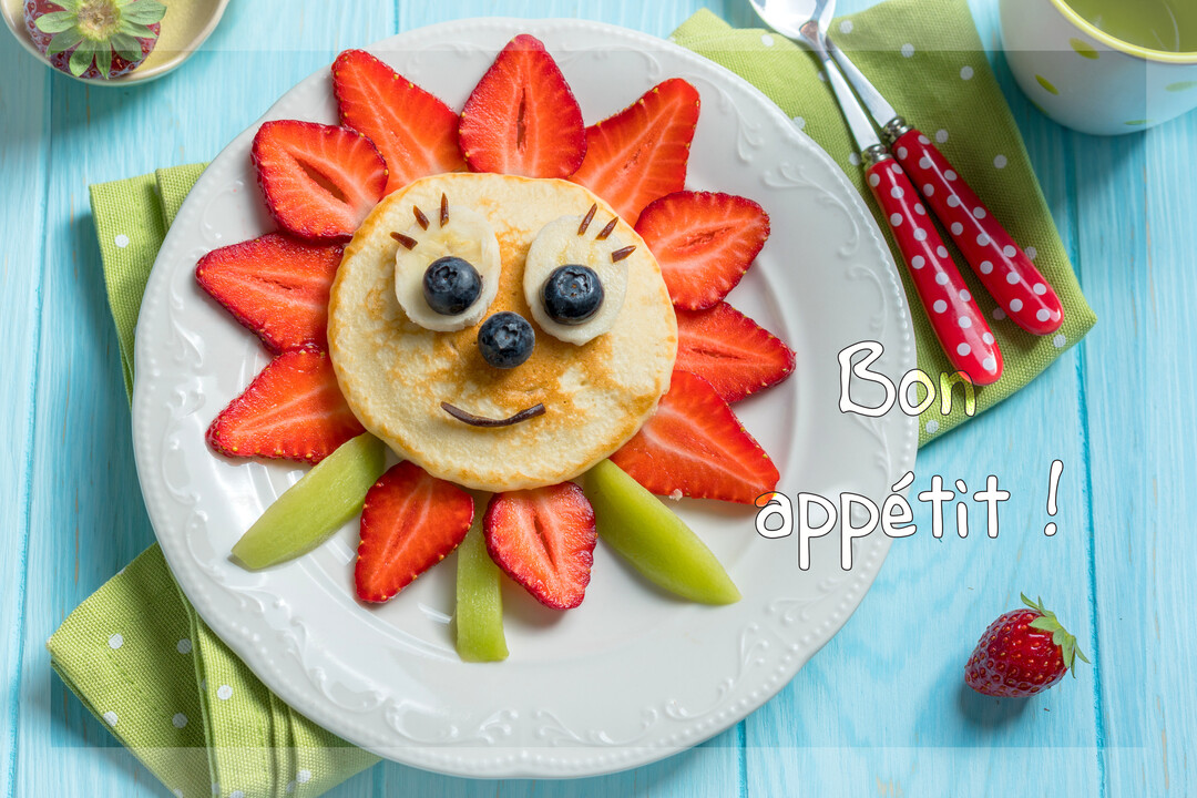 Une assiette blanche contenant un pancake décoré de fruits pour ressembler à un soleil souriant, sur une table en bois de couleur bleue.
