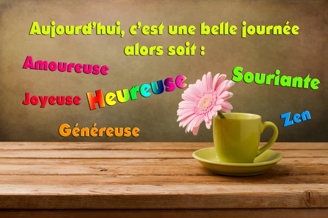 Une fleur de gerbera rose est placée dans une tasse verte sur une table en bois, avec un message coloré souhaitant une bonne journée.