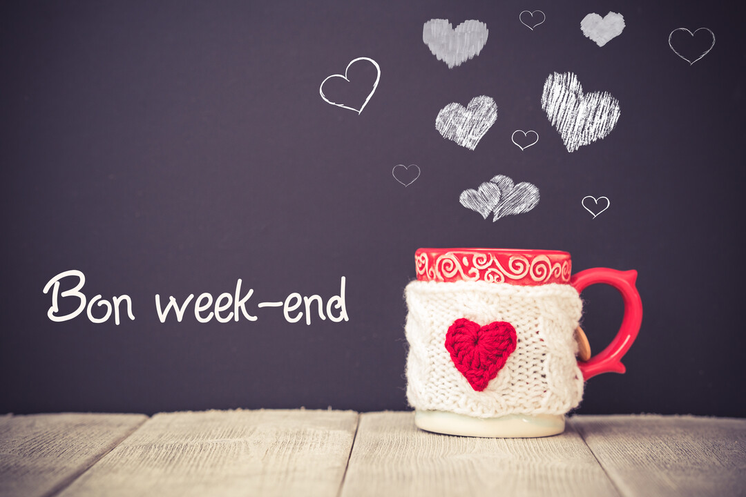 Une tasse de boisson chaude avec un cache-tasse en tricot orné d'un cœur rouge, posée sur une table en bois devant un tableau noir.