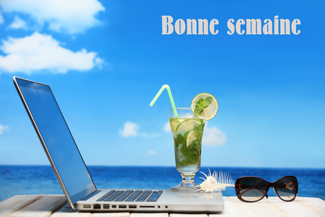 Un ordinateur portable est posé sur une table en bois au bord de la mer, à côté d'un cocktail, d'un coquillage et de lunettes de soleil.