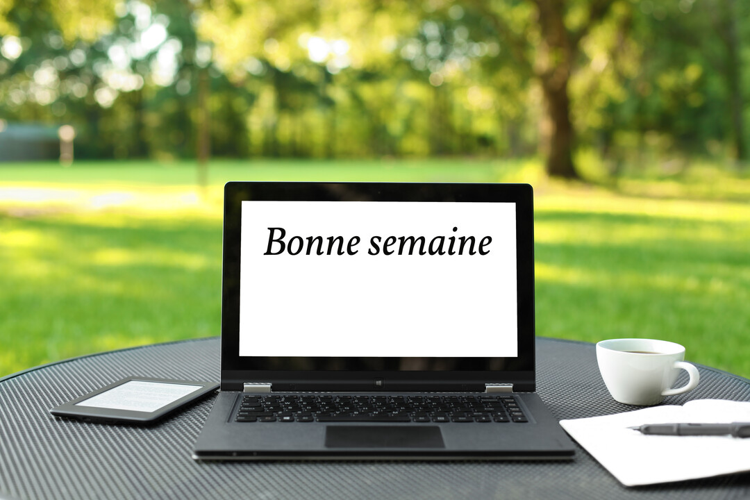 Un ordinateur portable affichant 'Bonne semaine' est posé sur une table dans un jardin, à côté d'une tasse de café et d'un carnet.