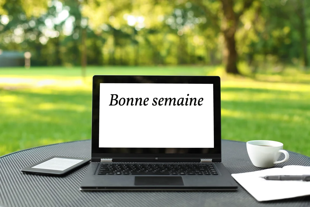 Un ordinateur portable affichant 'Bonne semaine' est posé sur une table dans un jardin, à côté d'une tasse de café et d'un carnet.