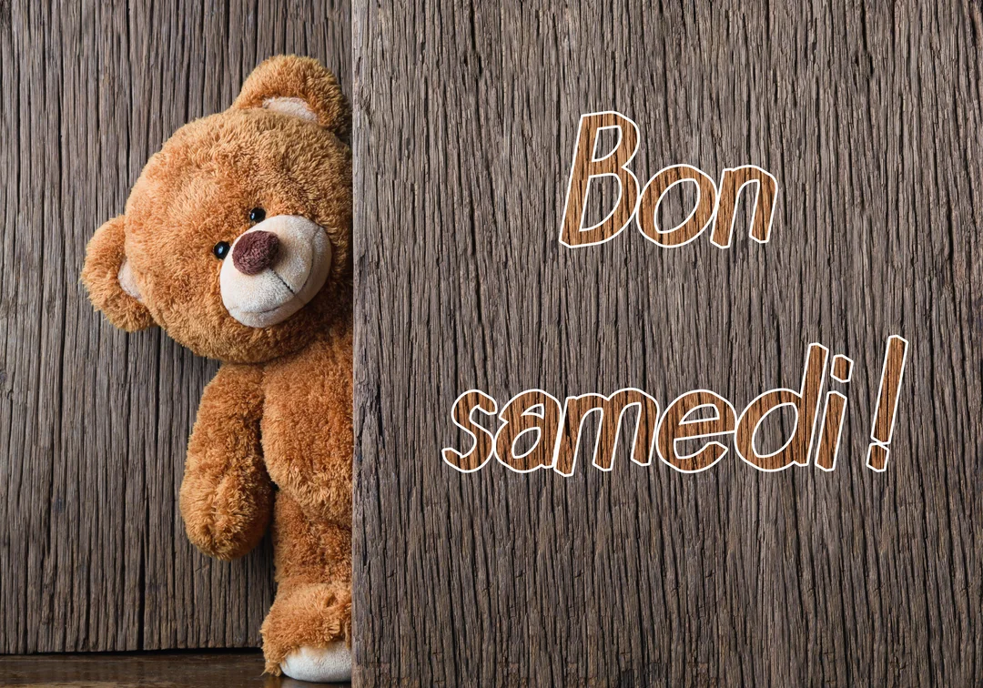 Un ours en peluche marron, avec un sourire malicieux, jette un coup d'œil derrière un mur en bois où est écrit 'Bon samedi'.