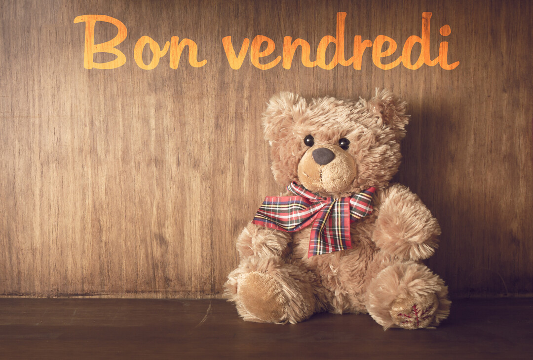 Un ours en peluche marron avec un nœud écossais est assis sur une surface en bois, devant un mur en bois où est inscrit 'Bon vendredi'.