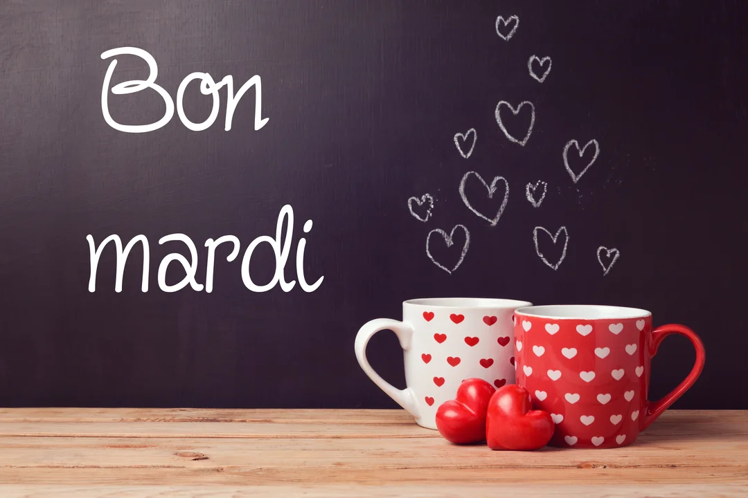 Deux tasses à café avec des motifs de cœurs sont posées sur une table en bois devant un tableau noir inscrit 'Bon mardi'.