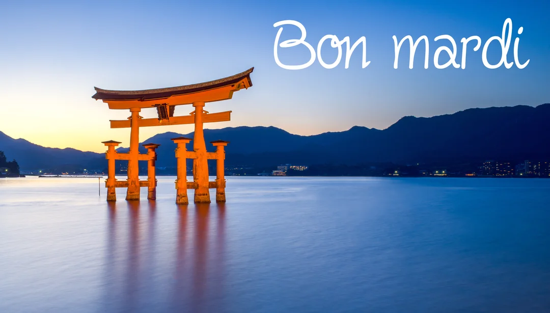 Un grand portail torii japonais orange se dresse dans l'eau paisible, face à des montagnes sous un ciel crépusculaire bleu et orangé.