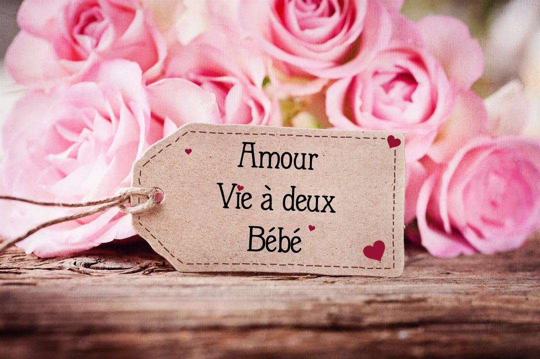 Une étiquette en papier kraft avec les mots 'Amour, Vie à deux, Bébé' repose sur une surface en bois devant un bouquet de roses roses.