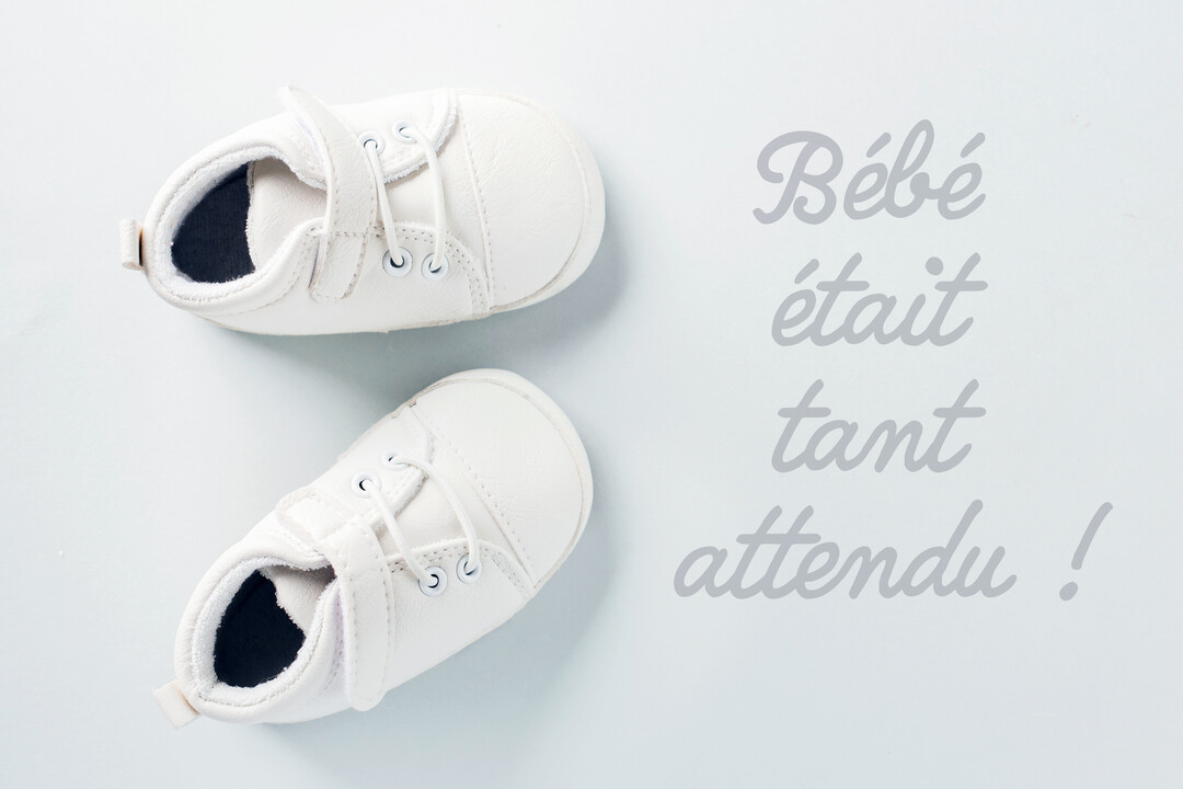 Vue de dessus d'une paire de baskets blanches pour nouveau-né posée sur un fond bleu-gris pâle à côté d'un message manuscrit.