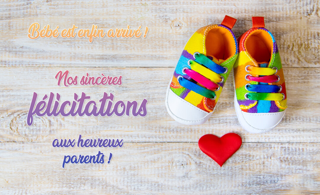 Sur un fond de planches de bois clair, une paire de petites baskets pour bébé aux couleurs vives est posée à côté d'un cœur rouge satiné.