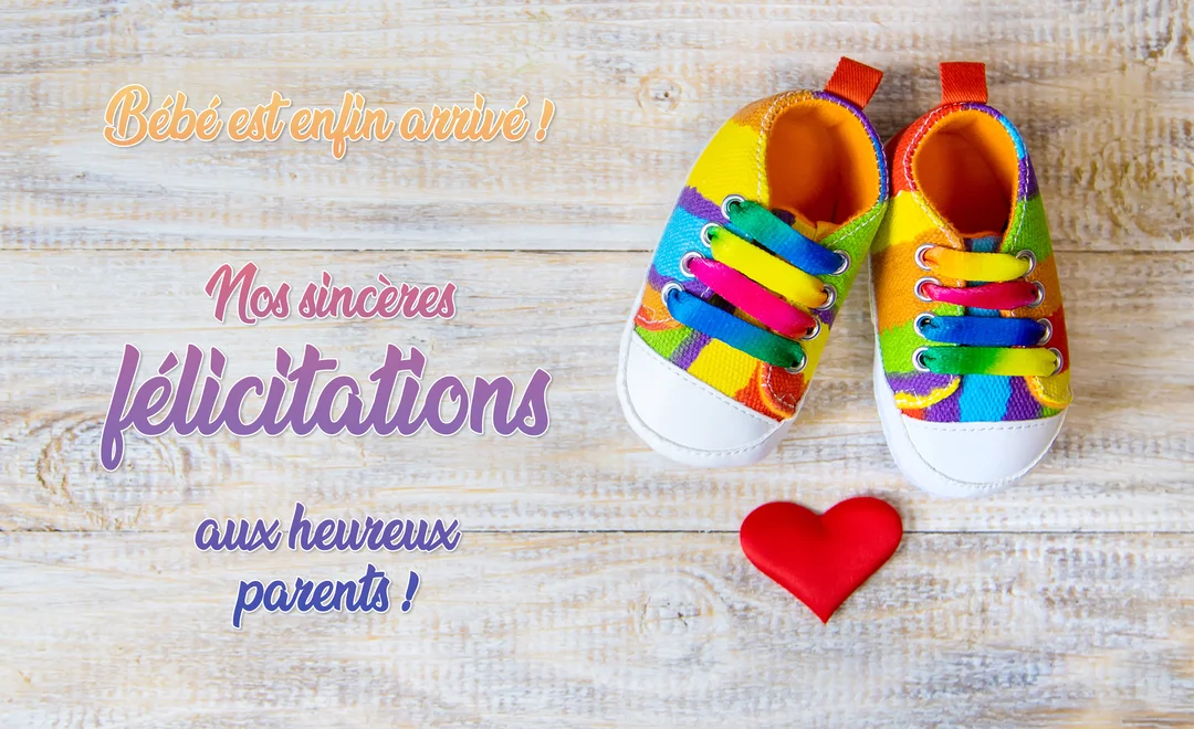 Sur un fond de planches de bois clair, une paire de petites baskets pour bébé aux couleurs vives est posée à côté d'un cœur rouge satiné.