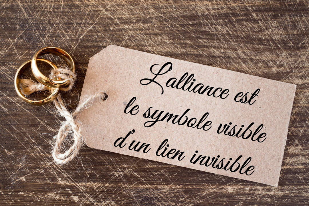 Deux alliances dorées sont liées par une ficelle à une étiquette rustique portant une citation poétique, le tout reposant sur une surface en bois sombre.