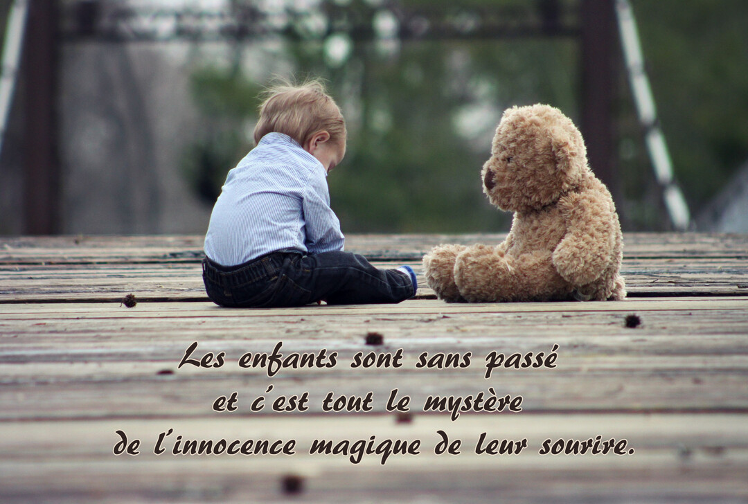 Un jeune garçon blond est assis sur un pont en bois face à son ours en peluche, dans une scène douce et contemplative.