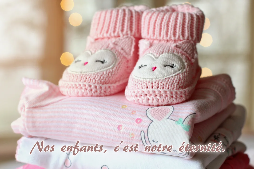 Une paire de chaussons roses pour bébé, ornés de visages d'animaux, est posée sur une pile de vêtements avec un fond lumineux.