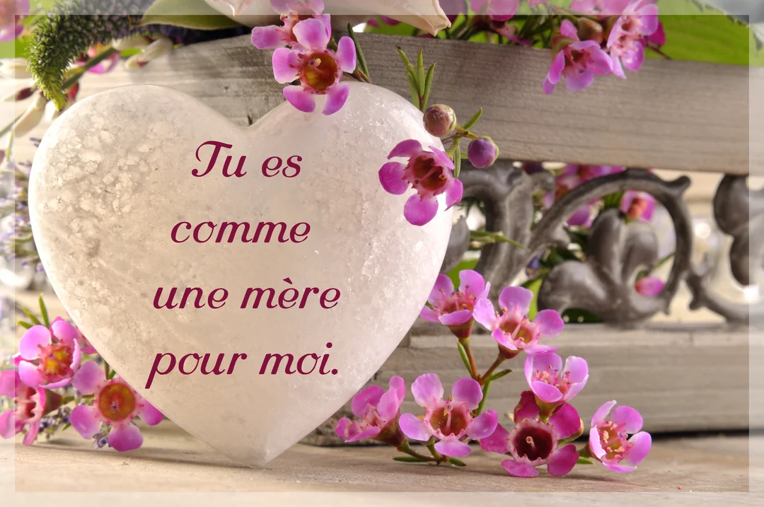 Un cœur blanc texturé portant une inscription est posé à côté de petites fleurs roses, sur un fond rustique en bois.