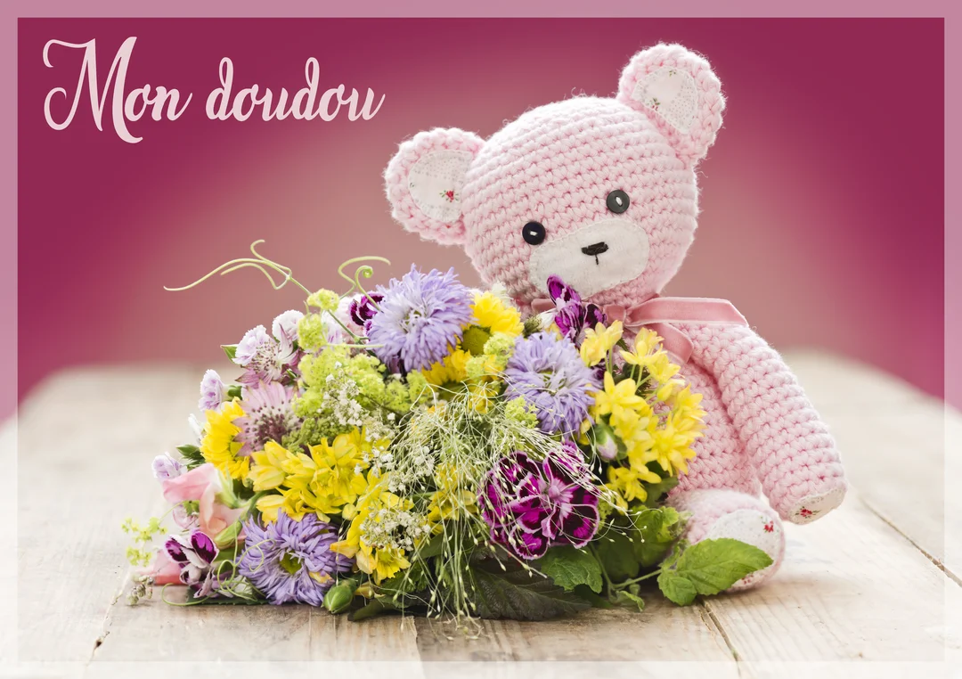 Un ours en peluche rose en crochet est assis sur une surface en bois à côté d'un bouquet de fleurs sauvages multicolores.