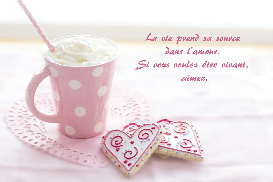 Une scène romantique et gourmande présente un mug rose à pois blancs avec chantilly et des biscuits en cœur sur un napperon assorti.