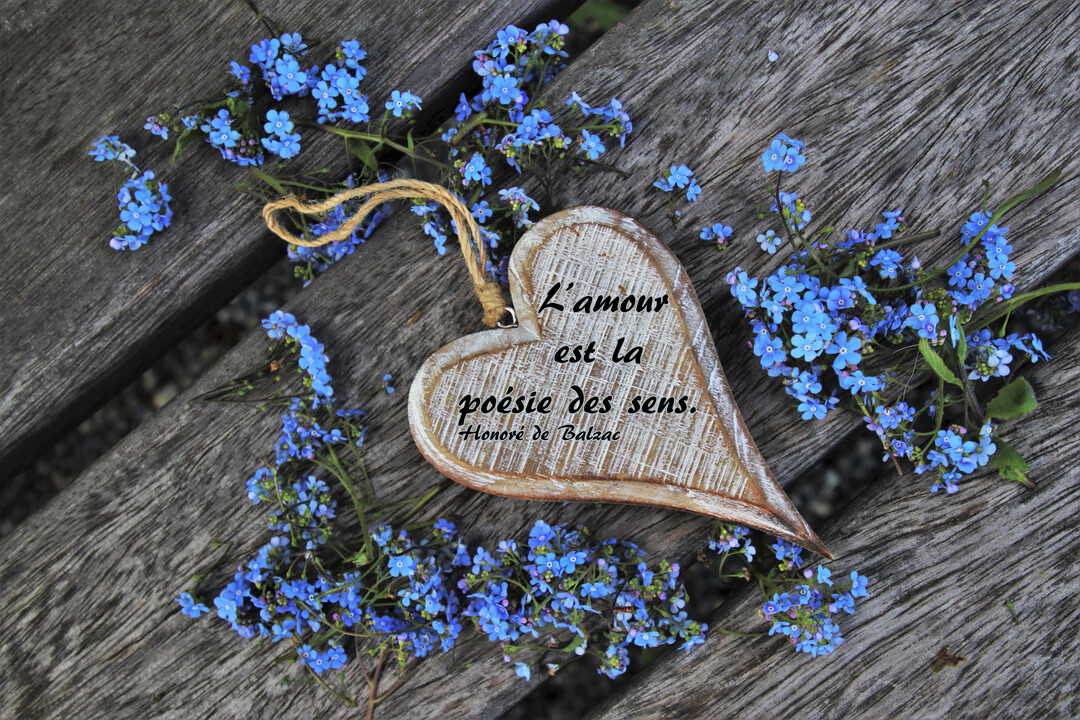 Un ornement en forme de cœur en bois est posé sur des lattes de bois, entouré de petites fleurs bleues de myosotis.