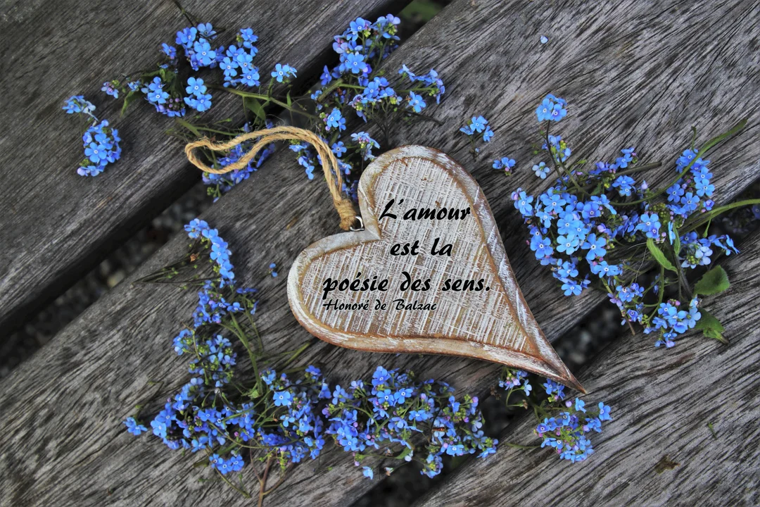 Un ornement en forme de cœur en bois est posé sur des lattes de bois, entouré de petites fleurs bleues de myosotis.